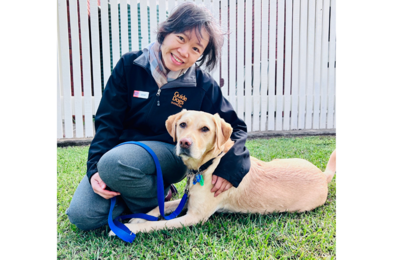 Clinical Psychologist, Dr. Sau Kuan Cheong (Janet) is sitting on the grass in front of a white fence. Jasmine, a yellow Labrador, lies by Janet's side.