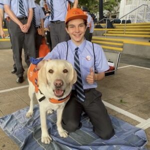 A Youth Ambassador from Brisbane Grammar school is sitting in a school yard next to a Yellow Labrador who is wearing an orange Guide Dogs branded coat. Both the Labrador and the boy appear to be smiling at the camera