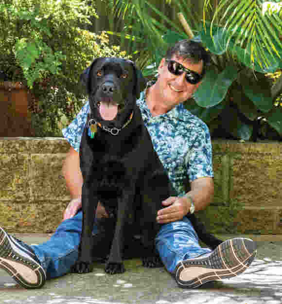 Image of Jason and Guide Dog Tyson sitting in front of a garden wall, smiling at the camera.