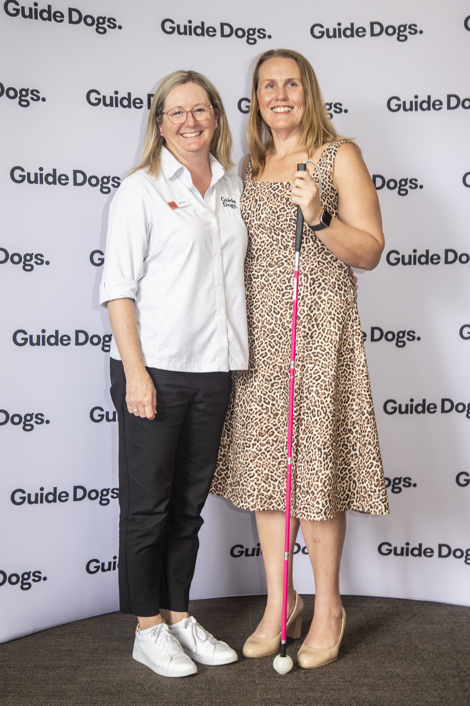 Karen (left) is standing next to one of her orientation and mobility Clients (right) who is holding their White Cane and smiling in front of a Guide Dogs media wall.