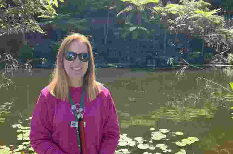 Sarah is standing in front of a pond surrounded by gardens and small waterfalls. Sarah is holding her White Cane and smiling at the camera