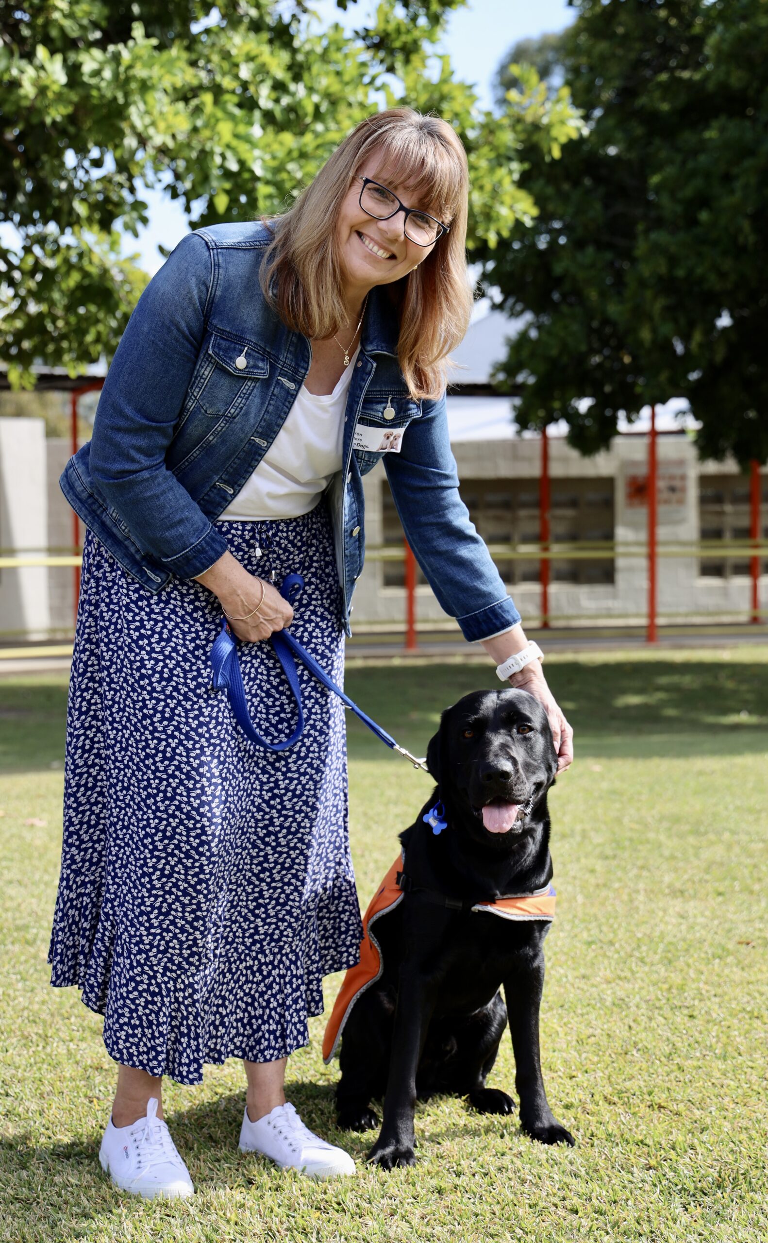 Sharon Masters is standing next to black Labrador in training, Davey who is wearing an orange Guide Dog in training coat. 