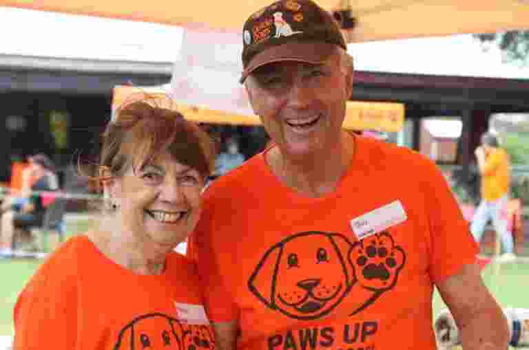 Image description: Wendy and Guy Mauger stand side by side dressed in orange Guide Dogs t-shirts at a busy volunteering event at Bald Hills.