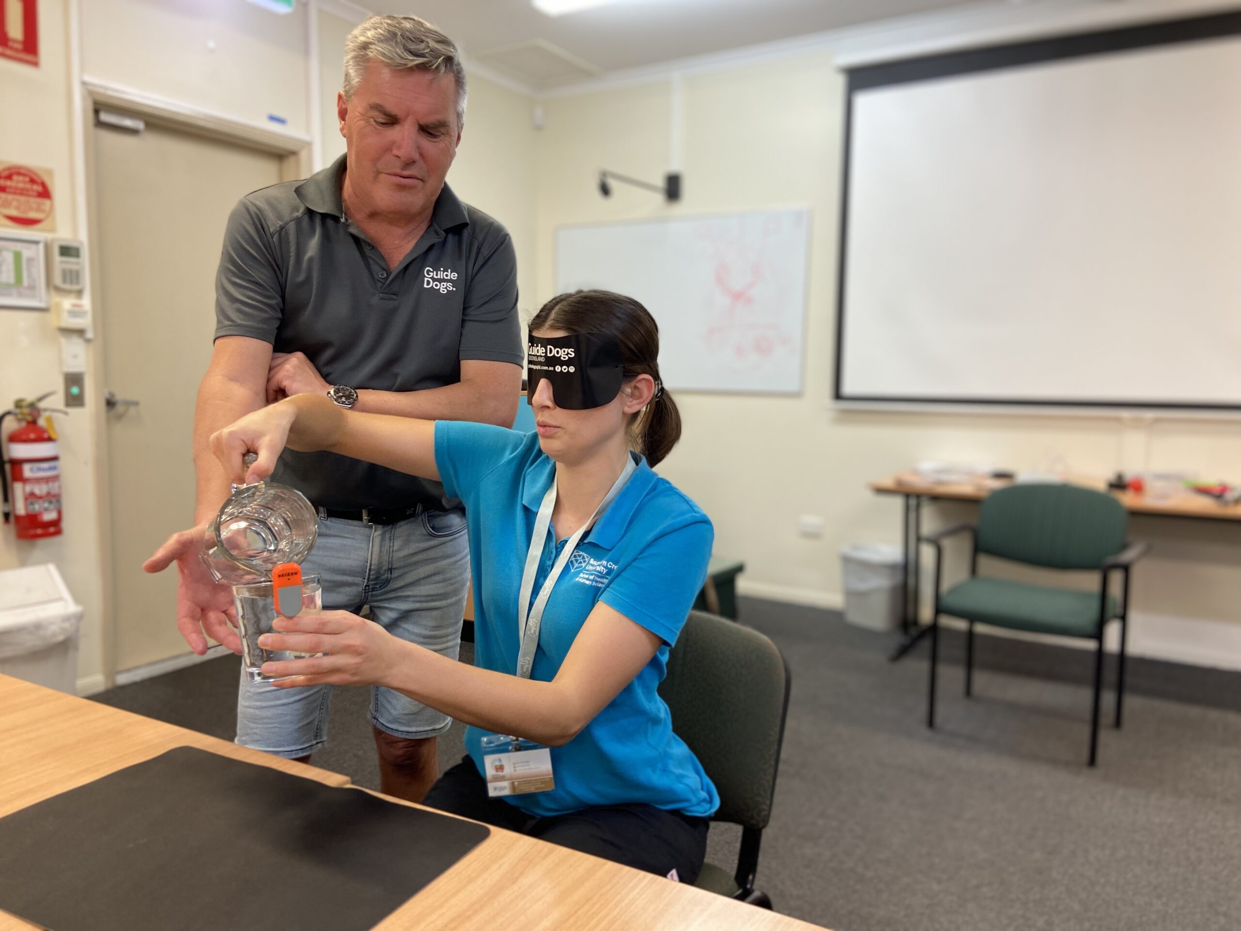 Guide Dogs Principal Advisor, Access and Engagement, Jeremy Hill, is pictured helping a participant from the Mount Isa workshop conduct a blindfolded exercise. The woman is seated at a table and is pouring a glass of water while wearing a blindfold.