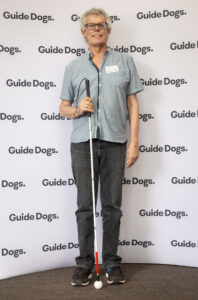 Occupational Therapy program graduate; George van der Heide smiling at the camera and standing in front of a Guide Dogs media wall with his white cane. 