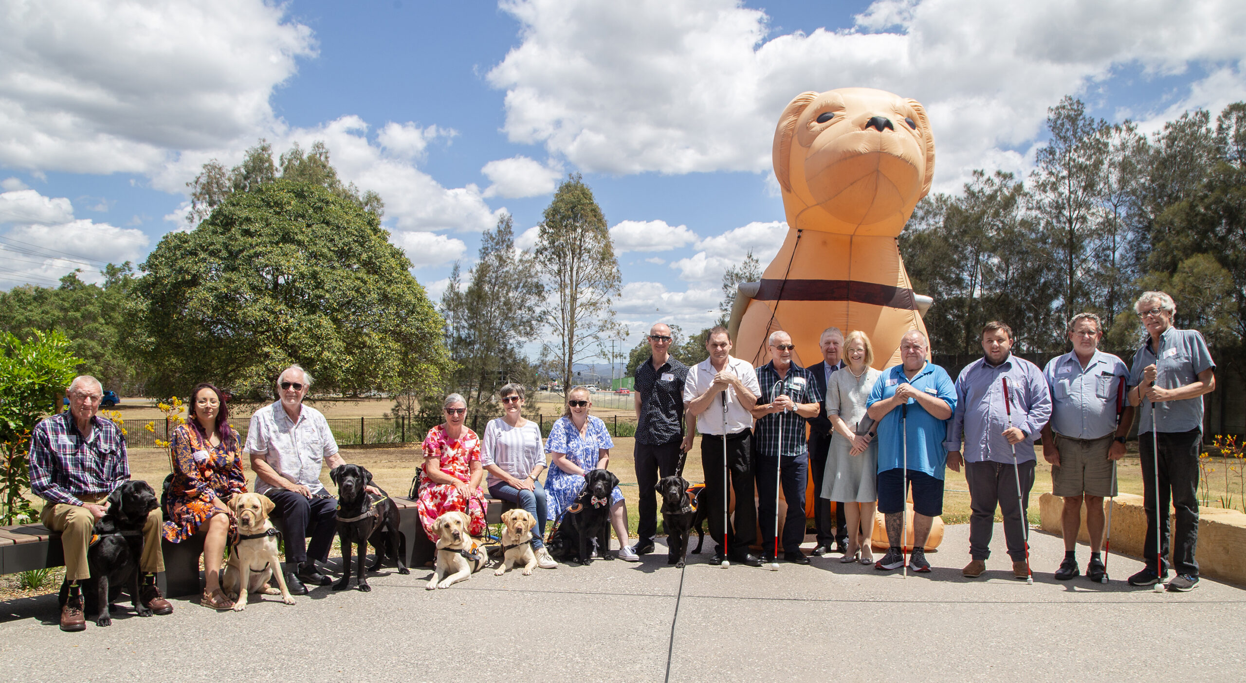 Group photo of attending Graduates with President of the Board Mr. Richard Anderson OAM and Her Excellency the Honourable Dr. Jeannette Young AC PSM, Governor of Queensland. Graduates are smiling at the camera and standing in front of inflatable Guide Dog Gulliver.