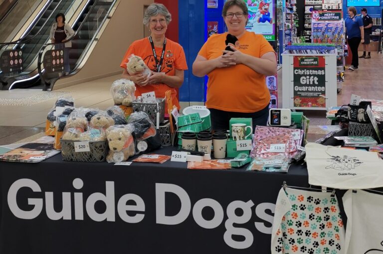 Two volunteers standing at a Guide Dogs merchandise stall, smiling.