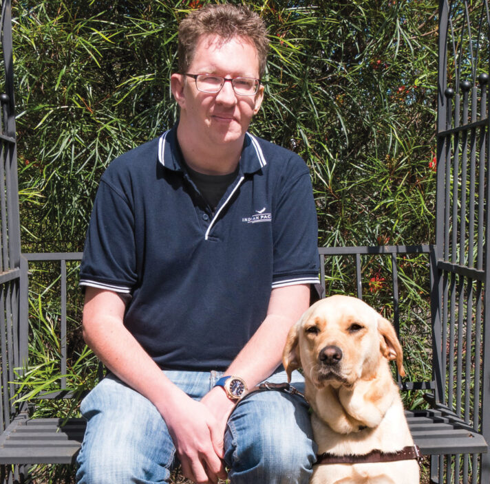 a man and a Guide Dog sitting by a bench, smiling.