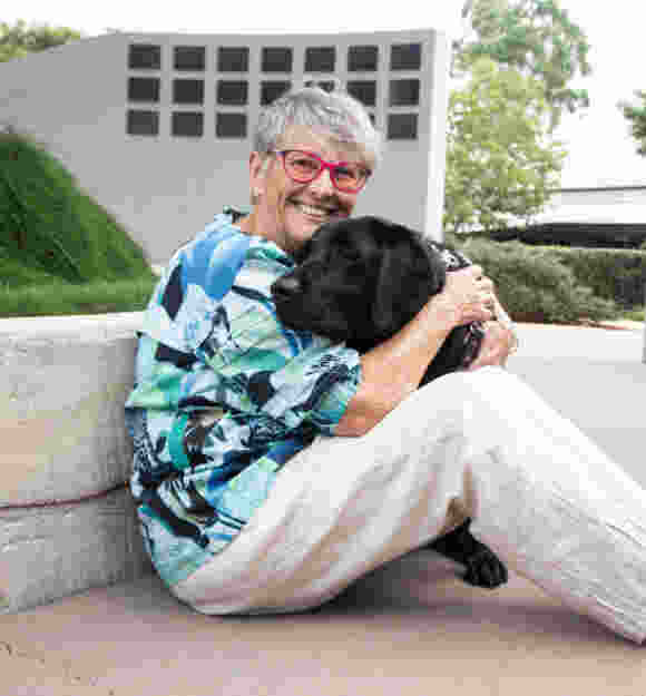 Sue is sitting on the ground in a colourful top hugging Guide Dog Vegas.