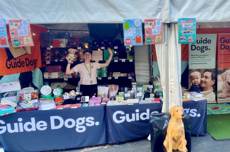 A Guide Dogs staff member stands behind a merchandise stall.