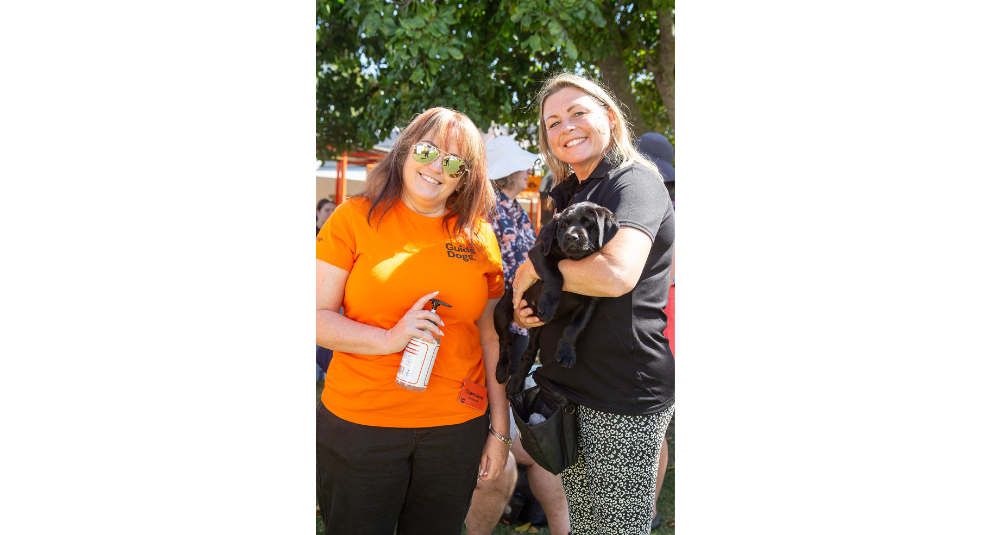A puppy trainer holding a black Labrador puppy and smiling at the camera. A volunteer in an orange shirt is standing beside them holding a bottle of hand sanitiser and smiling at the camera.