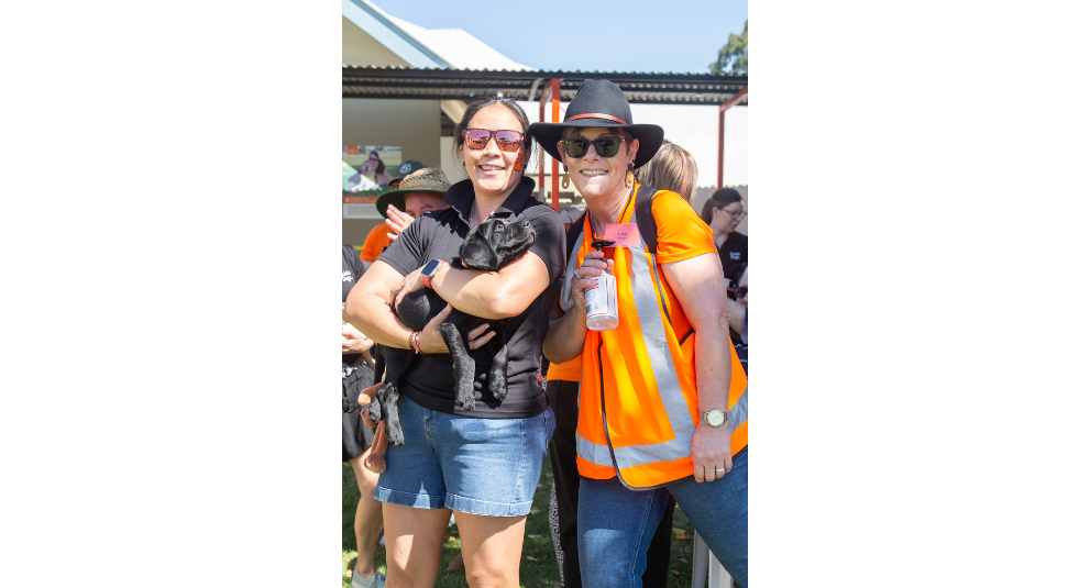 A puppy trainer holding a yellow Labrador puppy and smiling at the camera. A volunteer in an orange shirt is standing beside them holding a bottle of hand sanitiser and smiling at the camera.