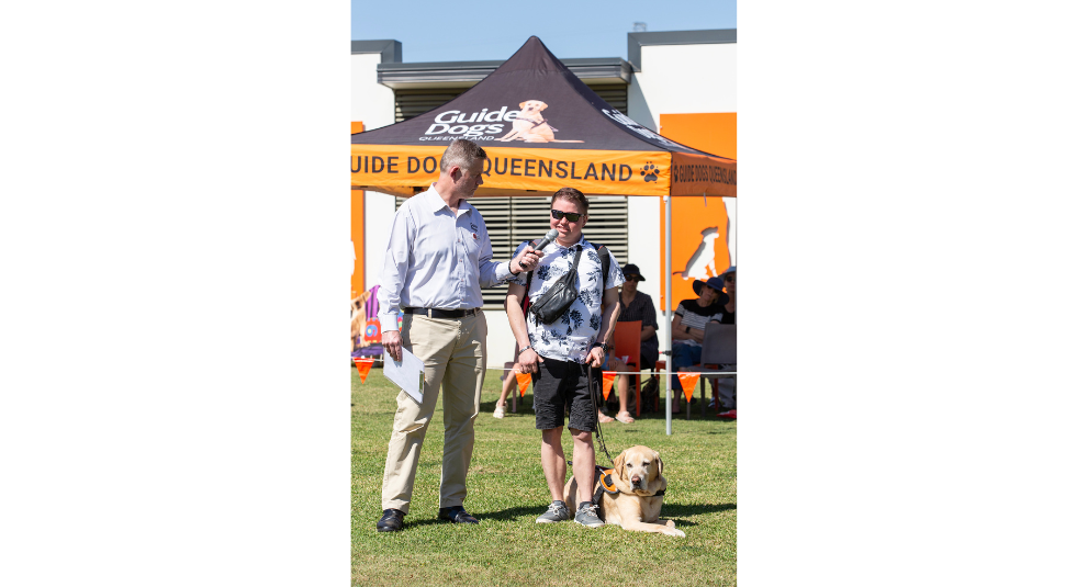 Guide Dogs CEO Jock Beveridge is holding a microphone while Client, Tyler, who is standing with his Guide Dog Fletcher beside Jock speaks to the crowd. They are facing towards the camera.