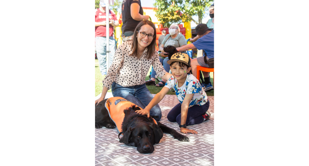 A woman and a boy are kneeling down patting a black Labrador in an orange training coat.
