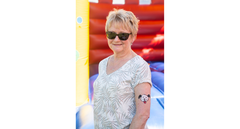 A woman wearing sunglasses standing in front of a jumping castle and smiling at the camera. There is a black and white dog face painted on her arm.