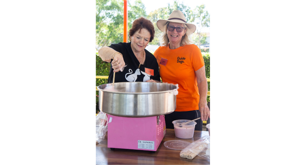 Two volunteers standing in front of a pink fairy floss machine. One woman is swirling the fairy floss onto a stick and the other is smiling at the camera.