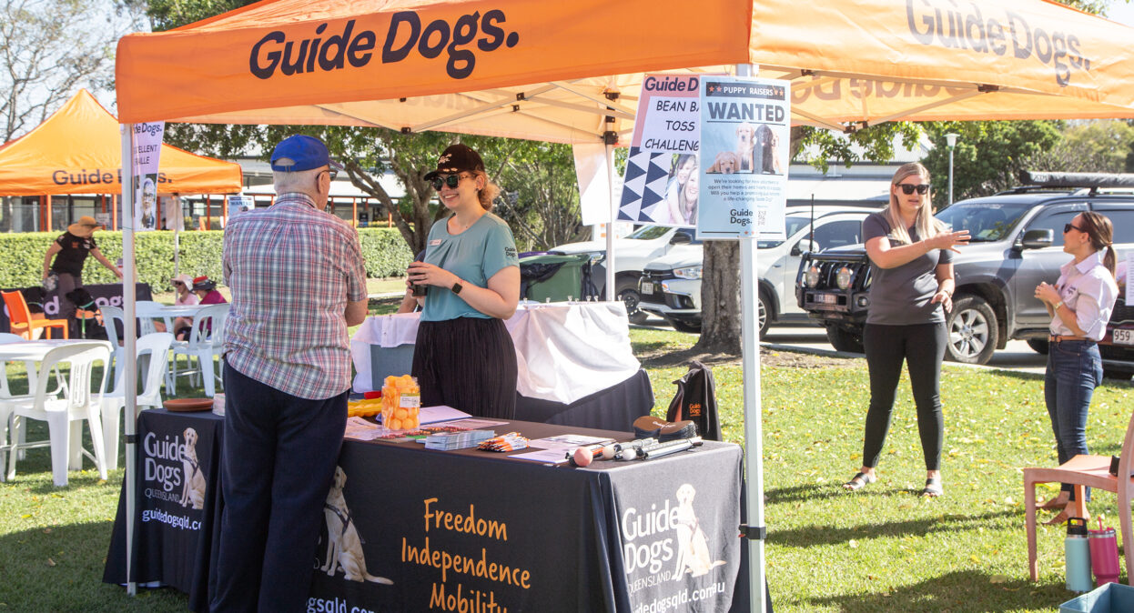 A man talking to a staff member at an outdoor Guide Dogs stall. The stall has an orange marquee with the Guide Dogs logo and a black tablecloth with Guide Dogs branding.