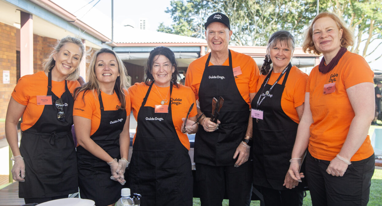 A group of volunteers in orange shirts and black aprons with the Guide Dogs logo are standing in a row and smiling at the camera.