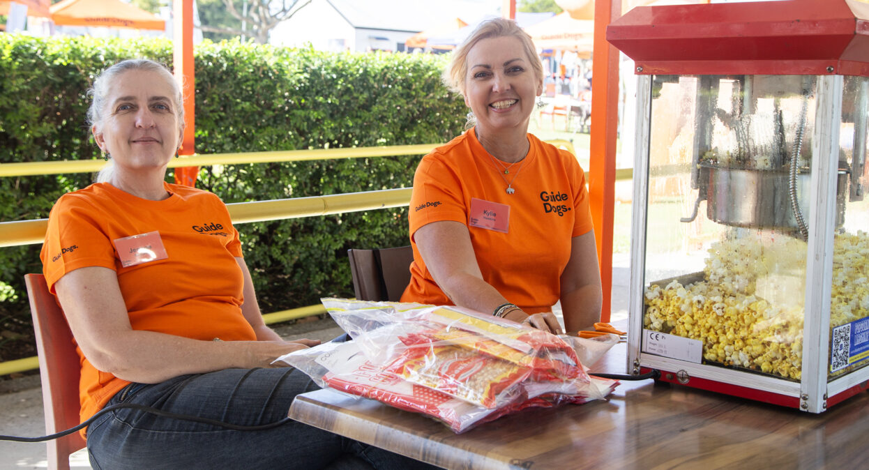 Two volunteers in orange shirts are sitting at a table behind a popcorn machine and looking at the camera.