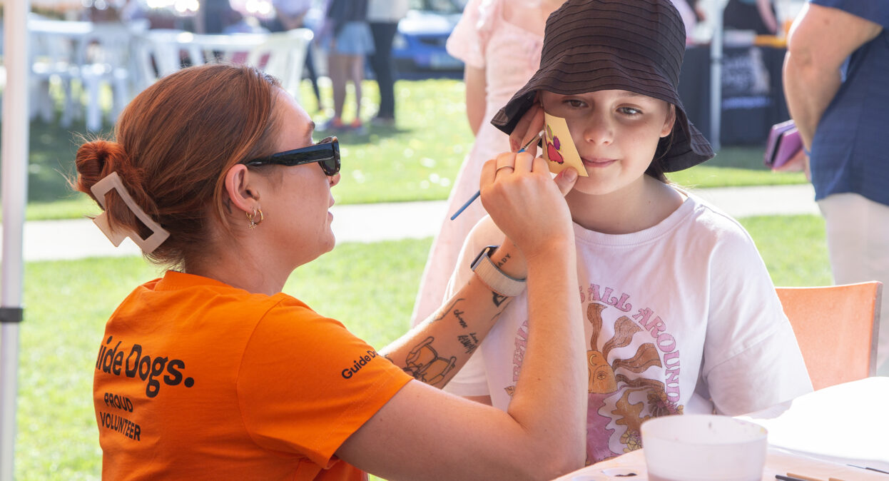 A volunteer in an orange shirt painting a paw print on a girl's cheek.