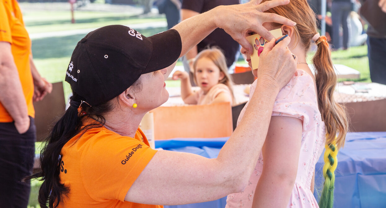 A volunteer in an orange shirt and a Guide Dogs hat painting a paw print on a girl's cheek.