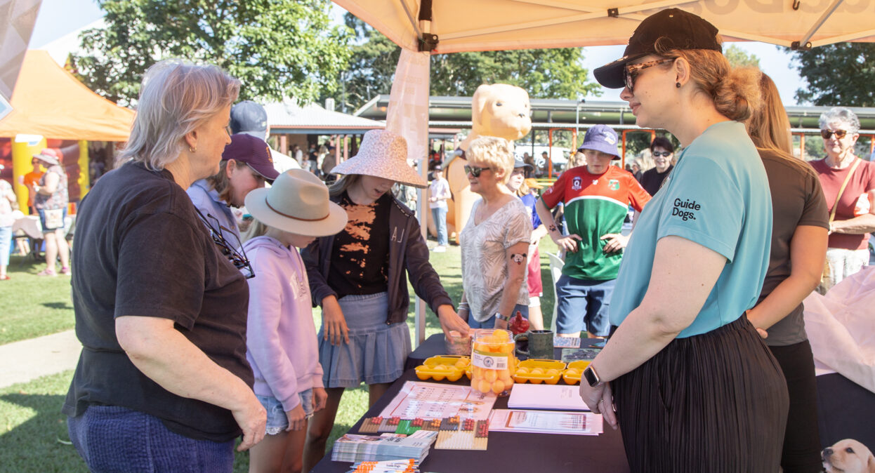 A group of people standing at a Guide Dogs stall where a staff member is standing behind a table. There are promotional pens, brochures and braille activities laid out on the table including ping pong balls and egg cartons.
