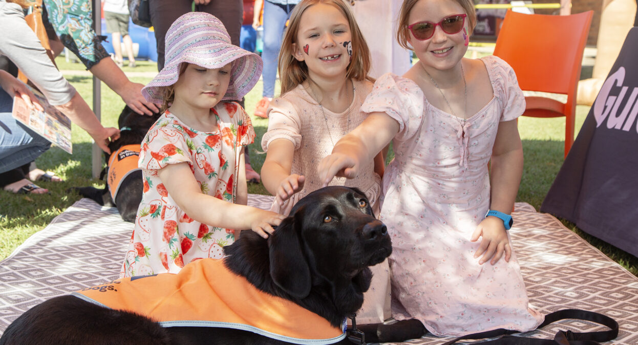 Three girls are kneeling behind a black Labrador in an orange training coat and patting the dog. The girls are smiling at the camera.