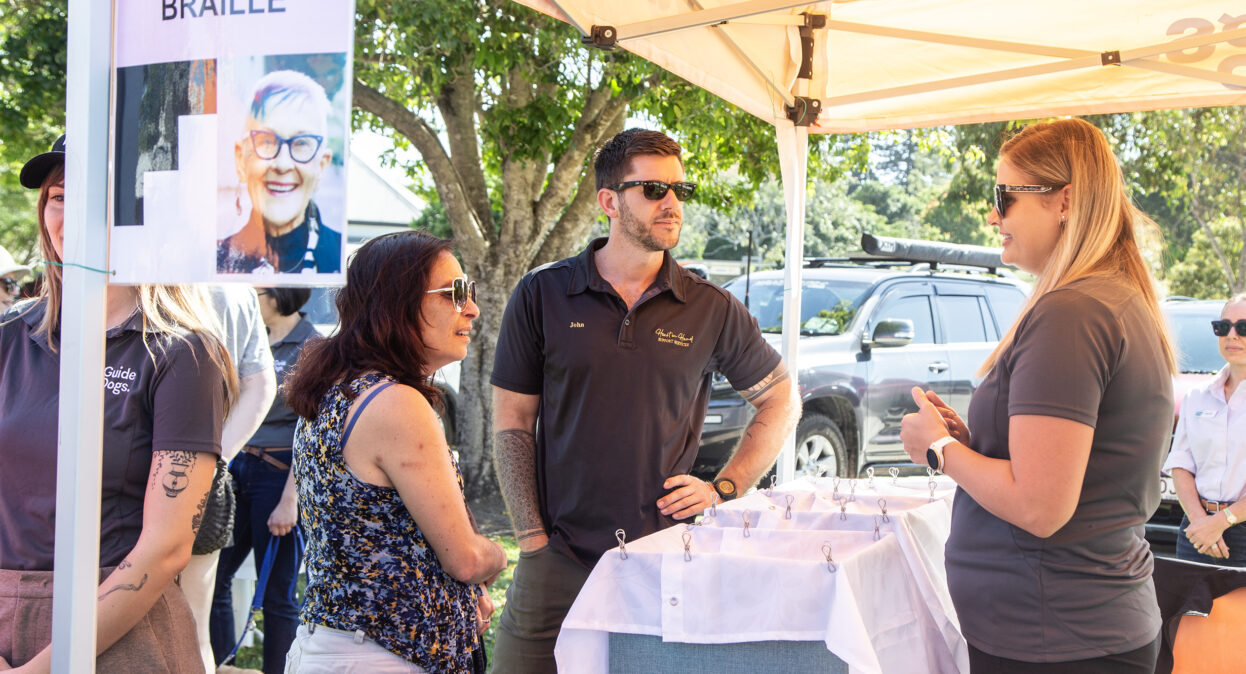 A main and a woman are standing at a stall speaking to a Guide Dog's staff member. There is a sign on the stall that reads 'EGGCELENT BRAILLE'.