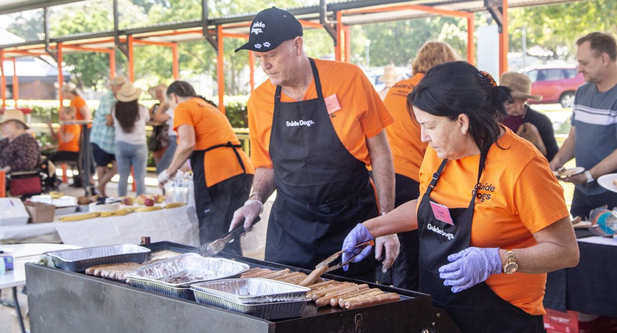 Volunteers in orange shirts and black aprons with the Guide Dogs logo are cooking sausages on a barbecue.