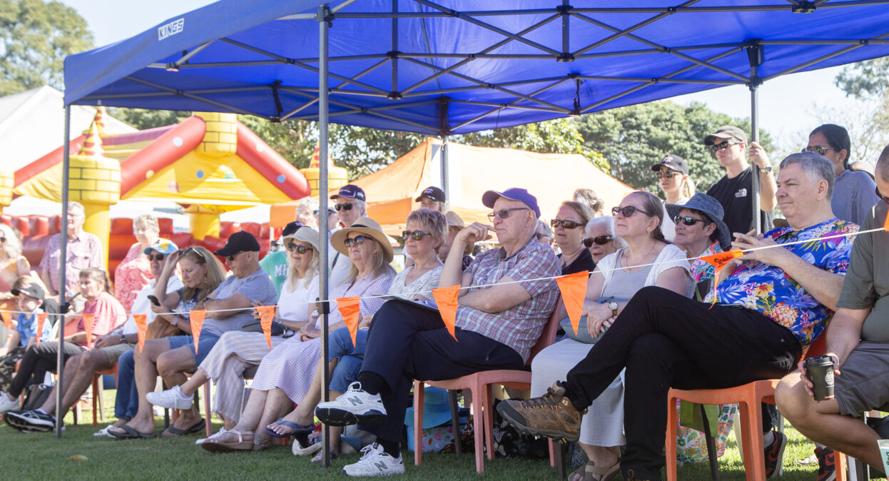 A crowd of spectators sitting and standing under a blue marquee next to a demonstration arena. There is a jumping castle in the background.