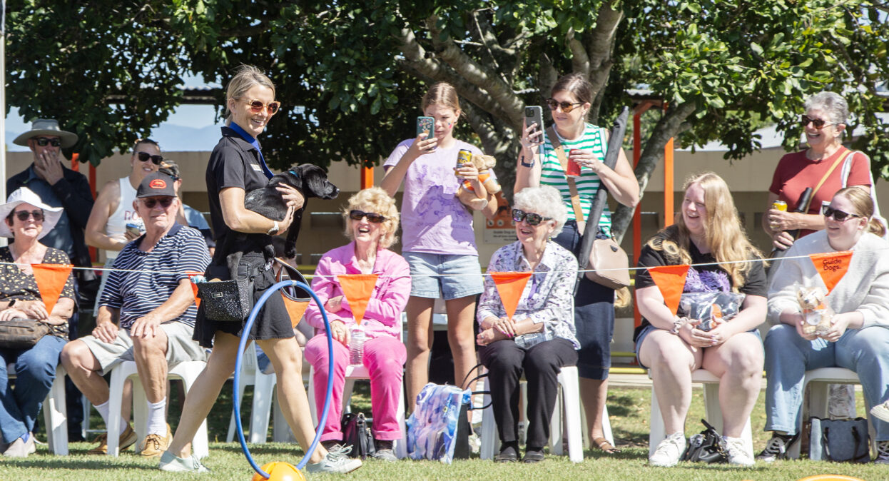 A puppy trainer carrying a black Labrador puppy around an arena as spectators watch. The spectators are looking at the puppy and smiling. Some people have their phone out and are taking photos.