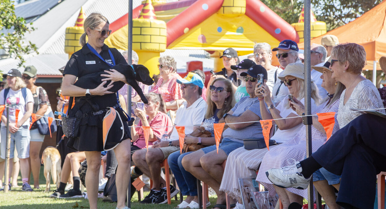 A puppy trainer carrying a black Labrador puppy around an arena as spectators watch. The spectators are looking at the puppy and smiling. Some people have their phone out and are taking photos.