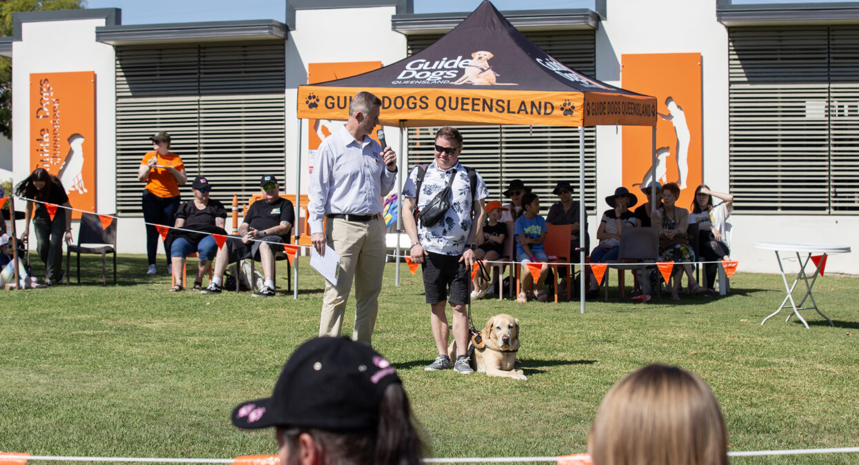 Guide Dogs CEO Jock Beveridge is holding a microphone and speaking to a Client, Tyler, who is standing with his Guide Dog Fletcher beside Jock. They are facing towards the camera.