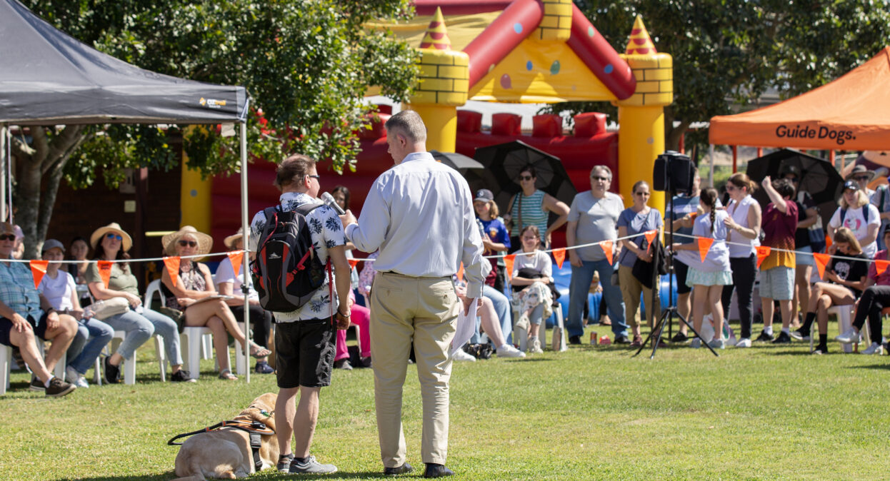 Guide Dogs CEO Jock Beveridge is holding a microphone and speaking to a Client, Tyler, who is standing with his Guide Dog Fletcher beside Jock. They are facing away from the camera.