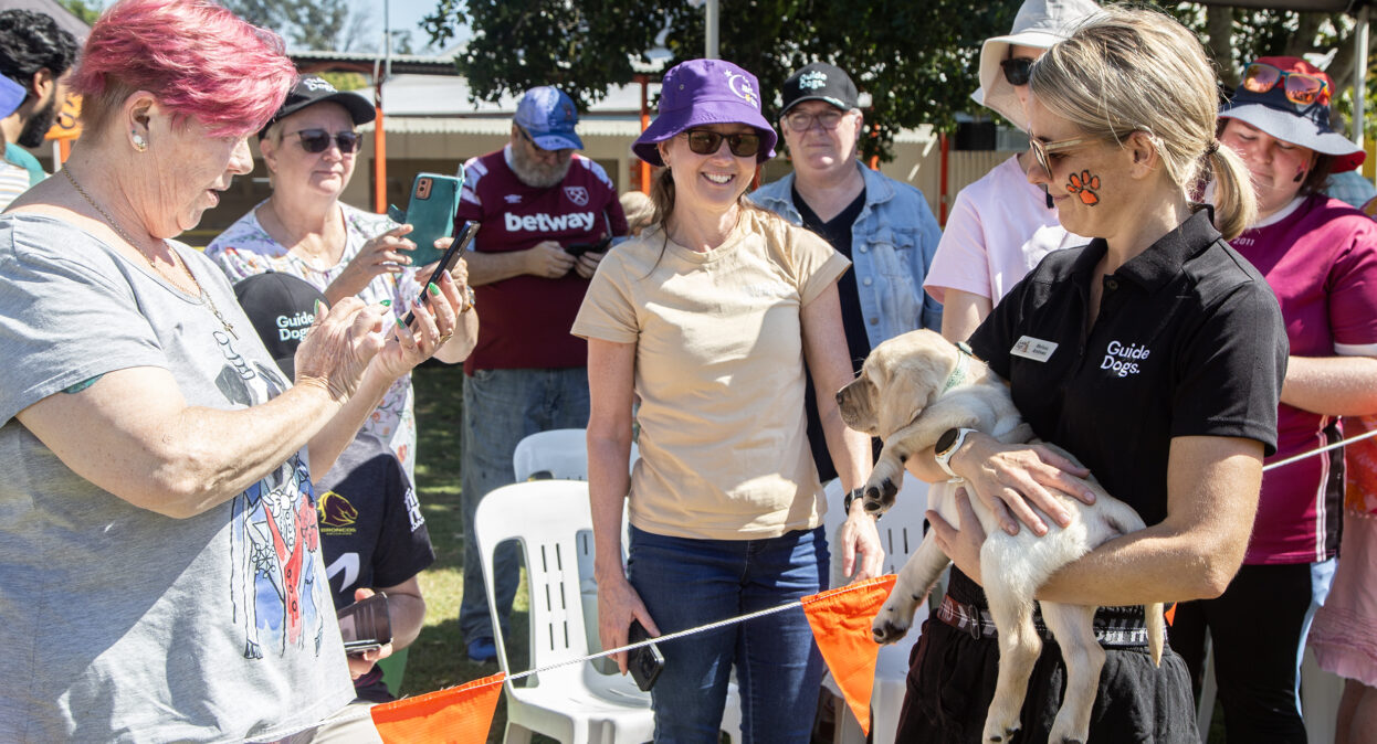 A puppy trainer holding a yellow Labrador puppy as a group of people take photos of the puppy on their phones.
