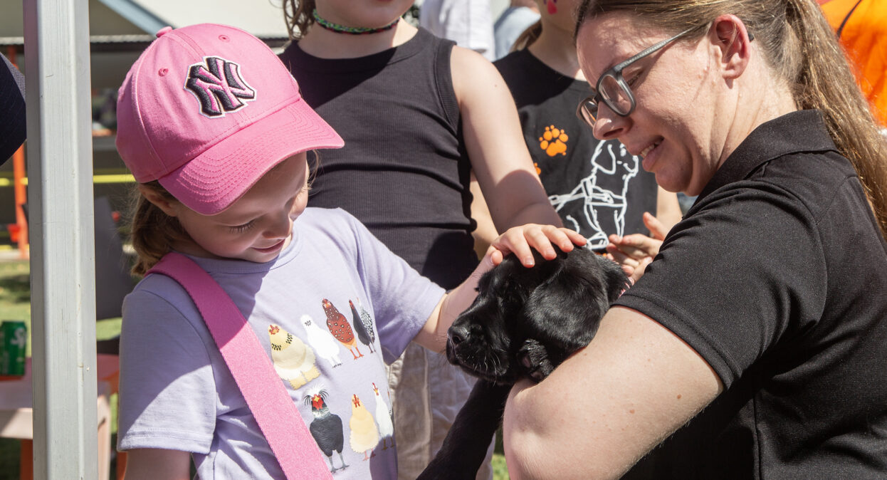A puppy trainer holding a black Labrador puppy as a young girl pats the puppy.