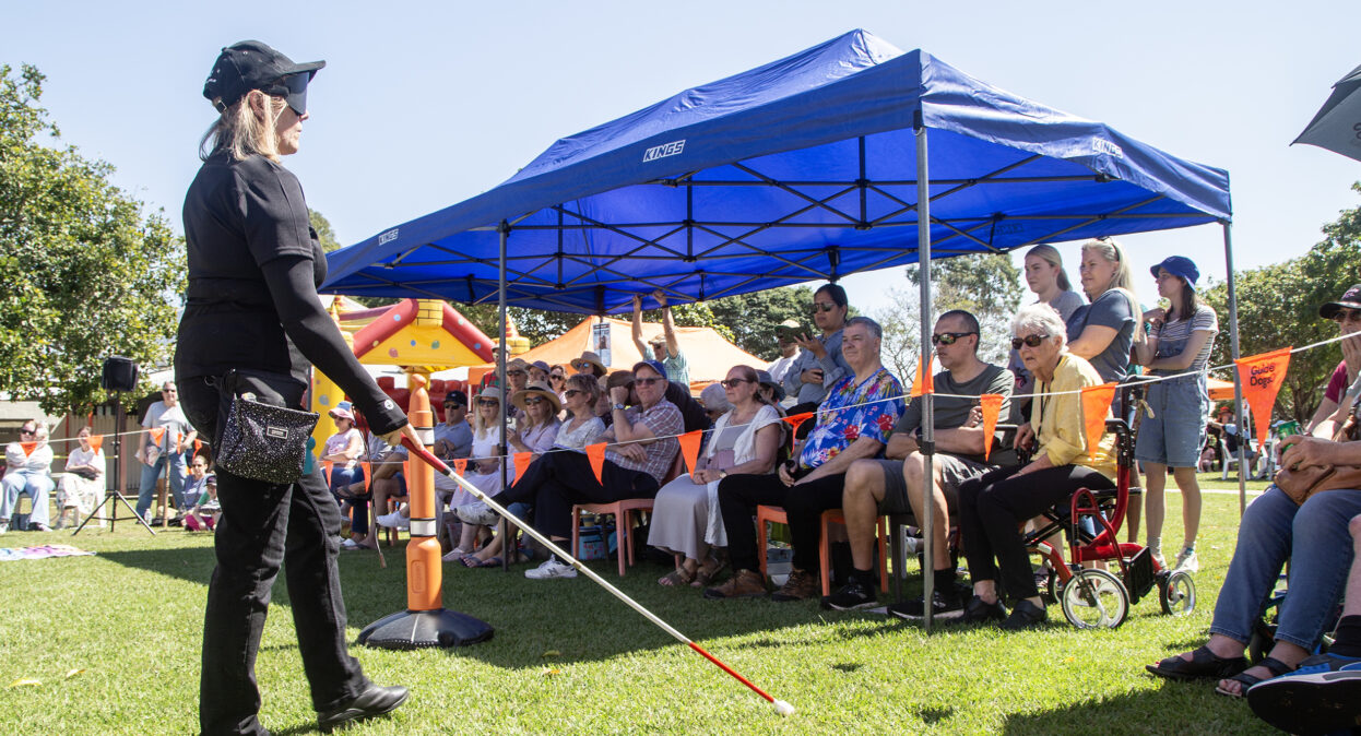 A Guide Dogs staff member under blindfold walking with a white cane in an arena with obstacle as a crowd of spectators watch the demonstration.