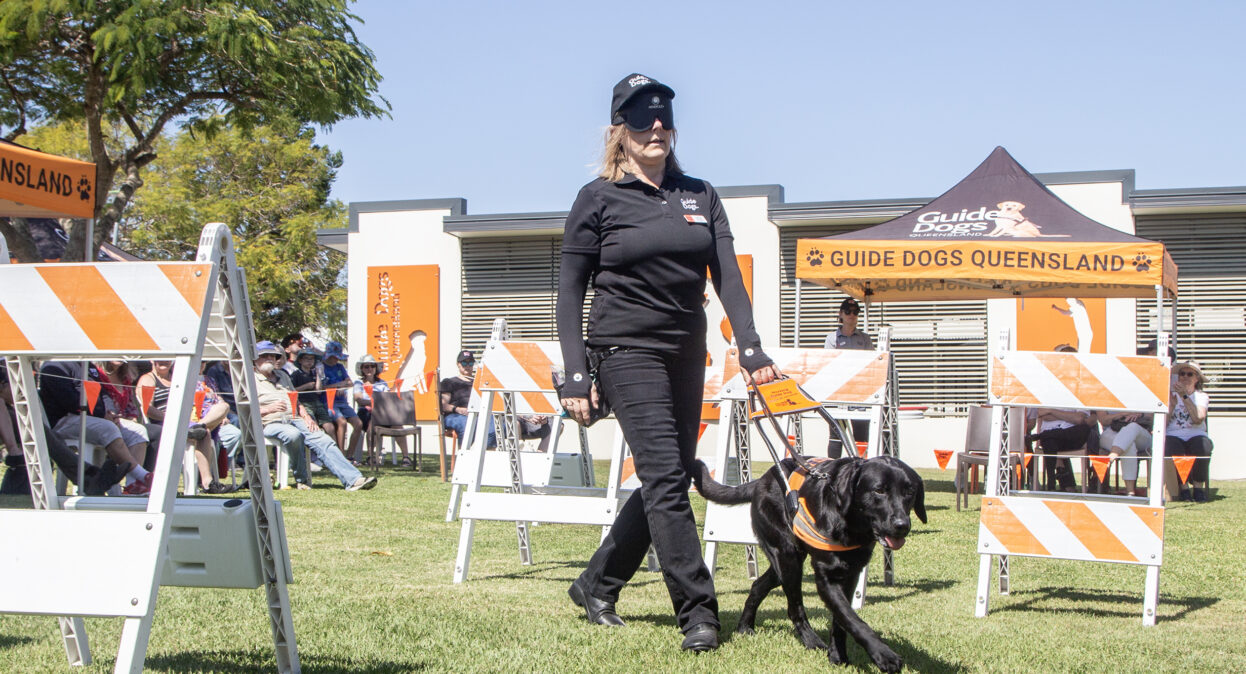 A Guide Dog Trainer walking under blindfold with a black Guide Dog in an arena with obstacles. A crowd of spectators in the background are watching the demonstration.