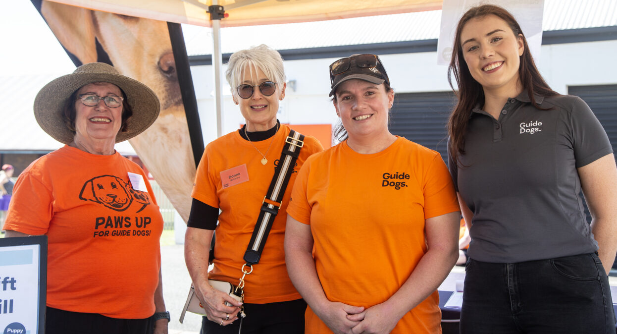 Three volunteers in orange Guide Dogs shirts stand beside a Guide Dogs staff member. They are all smiling at the camera.