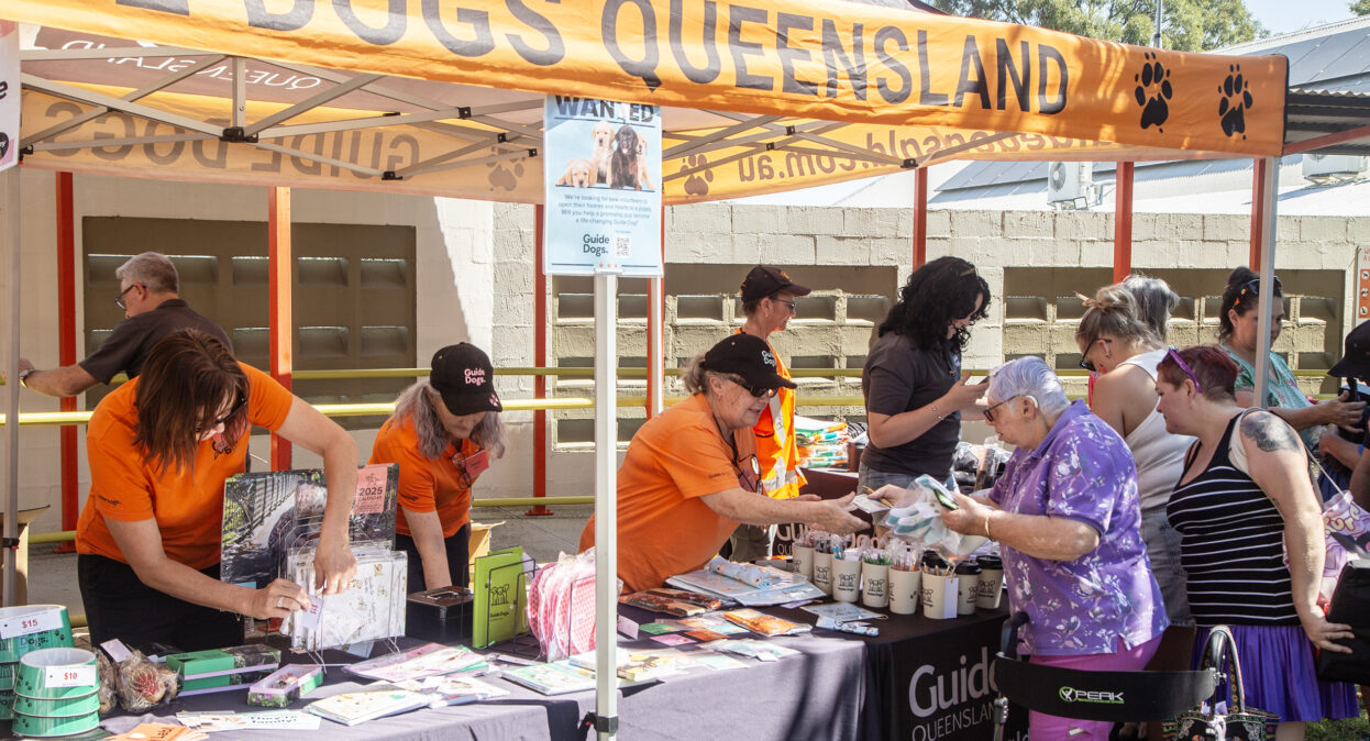 A Guide Dogs merchandise stall set up under Guide Dogs branded marquee. Volunteers in orange shirts are working at the stall and a group of customers are looking at items for sale.