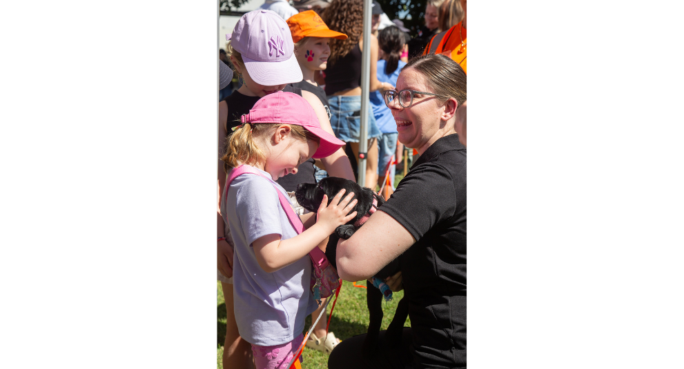 A puppy trainer holding a black Labrador puppy while a young girl pats the puppy.