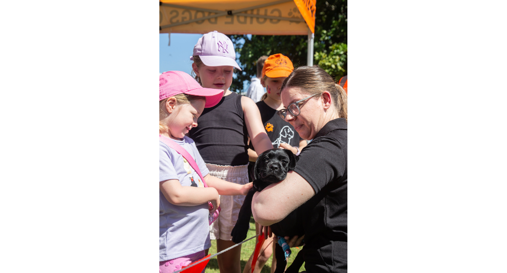 A puppy trainer holding a black Labrador puppy while a young girl pats the puppy.