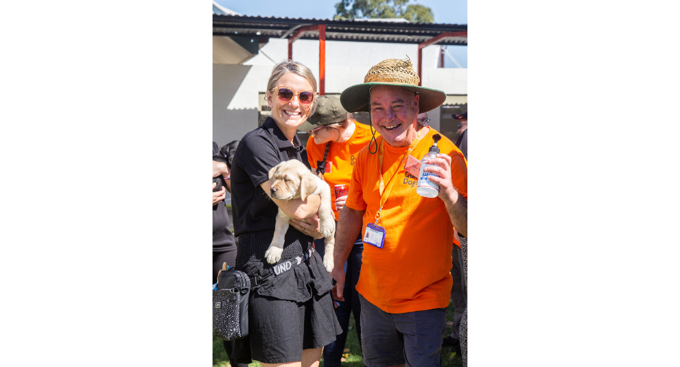 A puppy trainer holding a yellow Labrador puppy and smiling at the camera. A volunteer in an orange shirt is standing beside them holding a bottle of hand sanitiser and smiling at the camera.