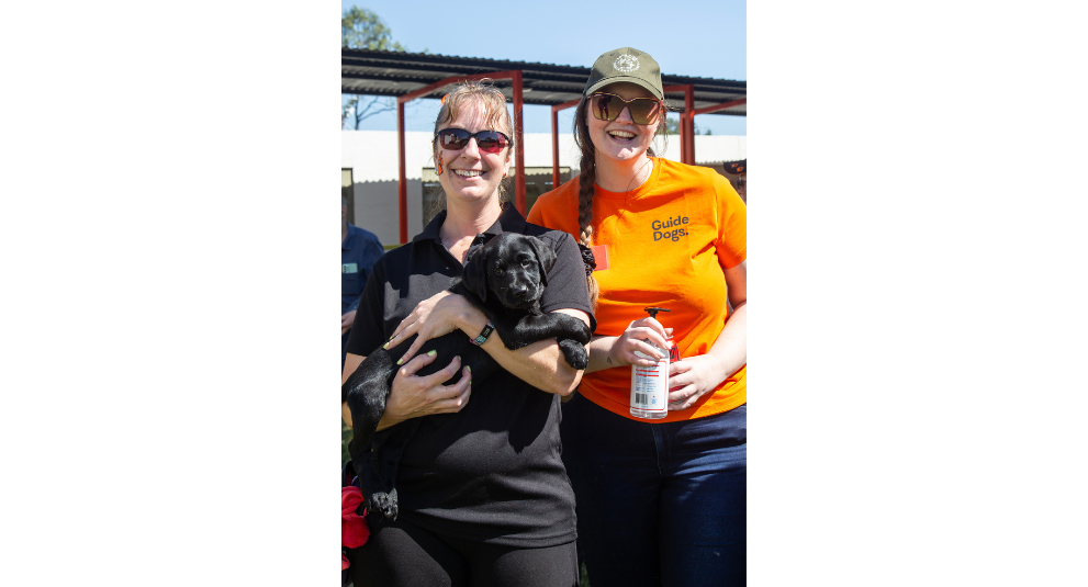 A puppy trainer holding a black Labrador puppy and smiling at the camera. A volunteer in an orange shirt is standing beside them holding a bottle of hand sanitiser and smiling at the camera.