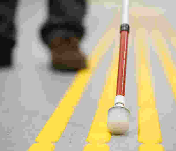 The end of a white cane is pictured up close on yellow tactile surface indicators.