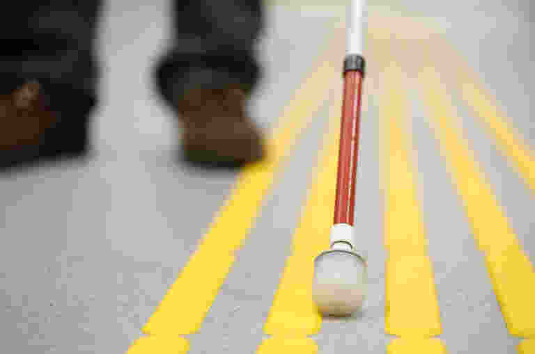 The end of a white cane is pictured up close on yellow tactile surface indicators.