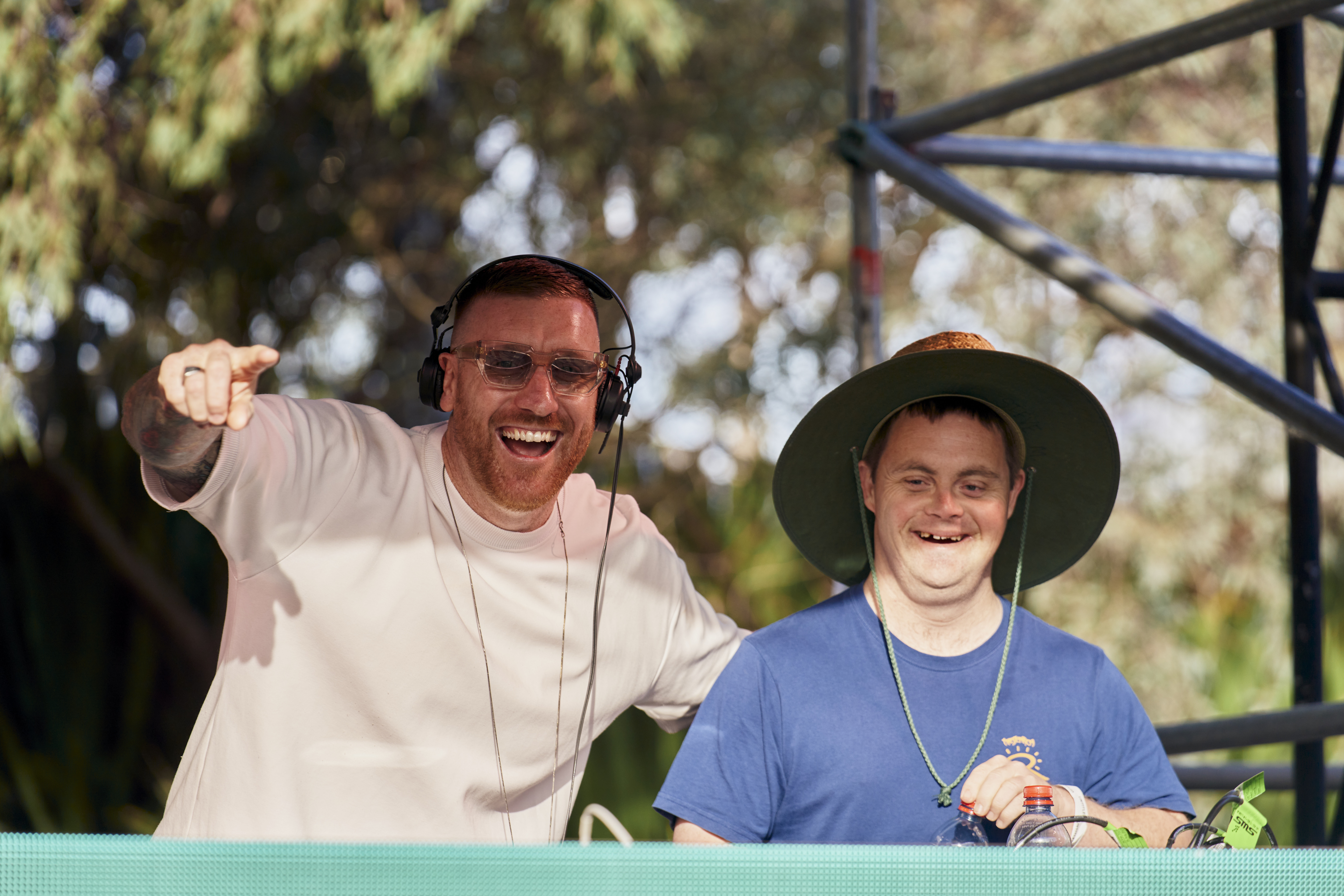 A DJ and a young man are pictured smiling to camera. The man on the left is a DJ wearing a headset and a white tshirt. The man on the right wears a blue shirt and a wide brimmed hat.