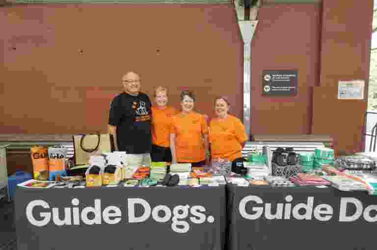 Four volunteers wearing Guide Dogs shirts are standing at a merchandise stall. The tables in front of them are covered in merchandise.