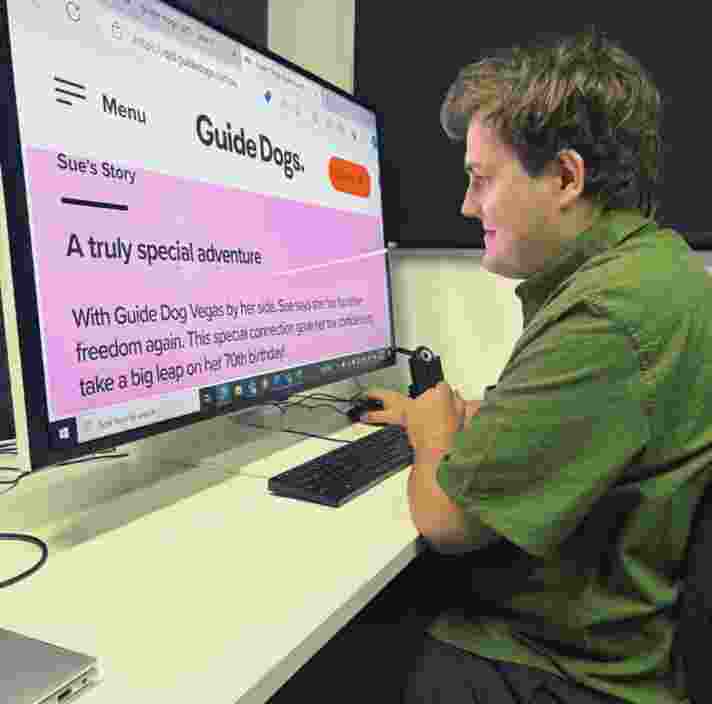 Image shows Brent, seated at a desk using a large computer screen with enlarged text displaying a Guide Dogs website. The screen shows a heading "A truly special adventure" and an article about Guide Dog Vegas. Brent is wearing a green shirt and is focused on the screen.
