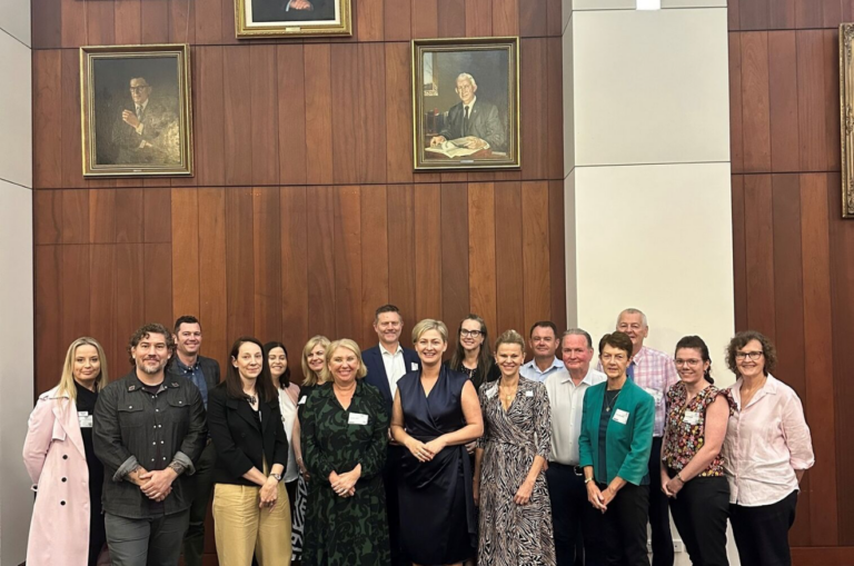 A group of people stand together smiling to the camera. They are in a room with high ceilings and images if politicians are on the walls above them.